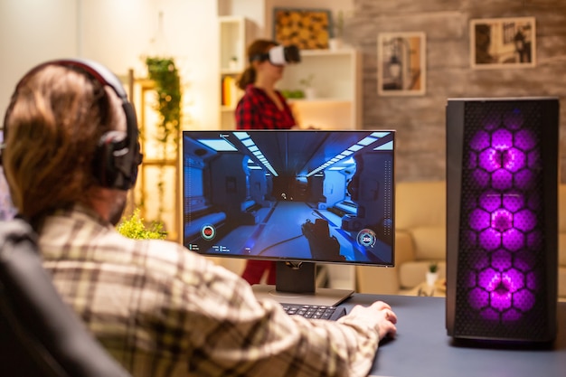 Vista traseira do videogame profissional jogando em um PC poderoso tarde da noite na sala de estar.