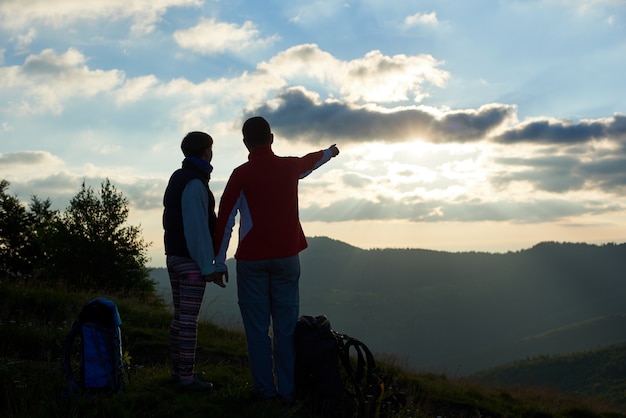 Vista traseira, dois turistas estão de pé no topo da montanha, segurando as mãos perto de mochilas contra o pano de fundo das montanhas e céu nublado ao pôr do sol. Homem mostrando a mão à distância