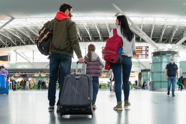 Vista traseira dos pais felizes carregando malas e segurando as mãos de sua filha enquanto vão ao longo do aeroporto antes do voo Conceito de viagem e viagem