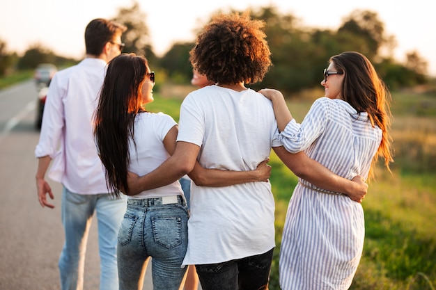 Foto vista traseira. jovem de cabelos escuros encaracolado abraçando duas meninas. eles conversam e caminham pela estrada. .