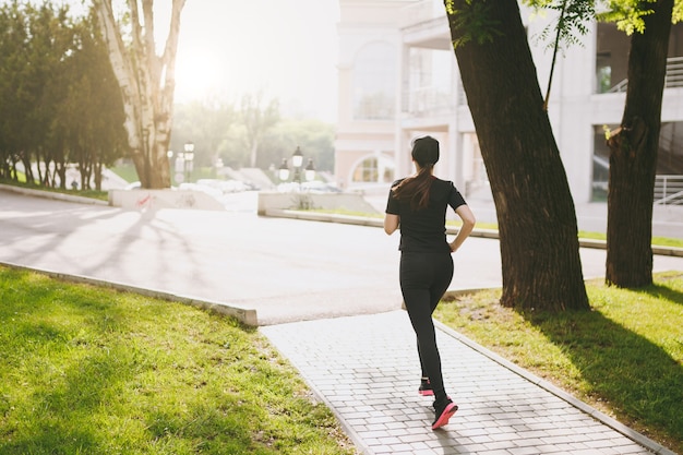 Vista traseira Jovem menina morena atlética de uniforme preto e boné treinando, fazendo exercícios de esporte e correndo, olhando direto no caminho no parque da cidade ao ar livre