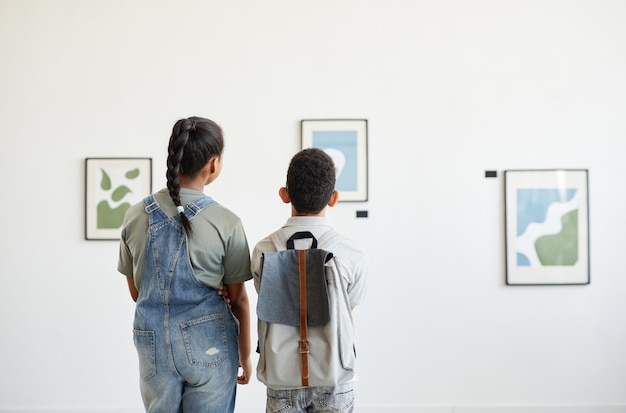 Vista traseira mínima de duas crianças visitando a galeria de arte e olhando pinturas abstratas modernas, copie o espaço