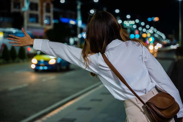 Vista traseira, mulher bonita sorrindo usando aplicativo de smartphone chamando com a mão para cima chamando táxi ao ar livre após o trabalho tardio, mulher de negócios asiática granizo acenando táxi de mão na estrada na rua da cidade à noite