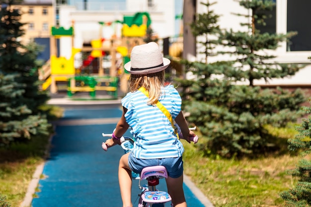 Vista traseira, uma adorável garotinha loira gosta de andar de bicicleta pequena pelo caminho no parque verde de verão