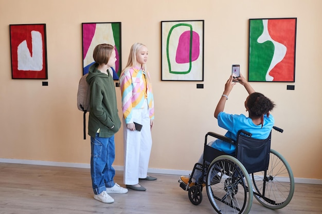 Vista trasera de un adolescente en silla de ruedas tomando fotos de cuadros en una galería de arte