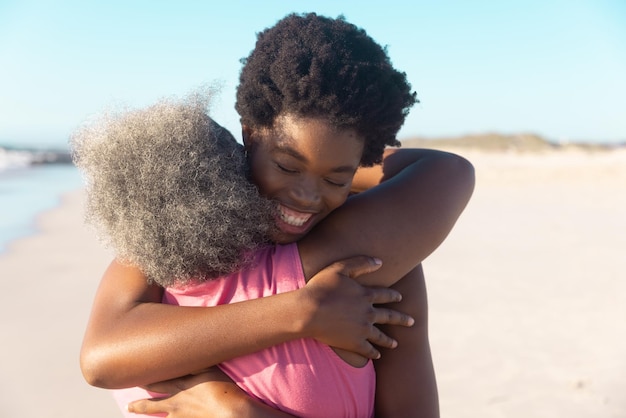 Vista trasera de una anciana afroamericana abrazando a su joven hija feliz en la playa bajo un cielo azul. Copiar espacio, inalterado, familia, verano, juntos, vacaciones, jubilación, disfrute y concepto de naturaleza.