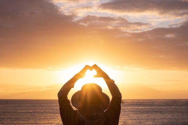 Vista trasera de una anciana caucásica al atardecer dorado de pie en la playa gesticulando en forma de corazón con las manos mirando el horizonte sobre el agua Relajada anciana de cabello blanco disfrutando de vacaciones en el mar