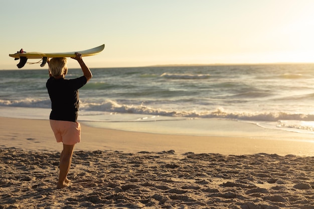 Vista trasera de una anciana caucásica en la playa al atardecer, sosteniendo una tabla de surf en la cabeza y mirando al mar