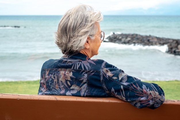 Vista trasera de una anciana sentada en un banco en la playa en un día de invierno disfrutando de vacaciones de tiempo libre o de jubilación Mujer de cabello gris a la moda con anteojos relajado frente al mar