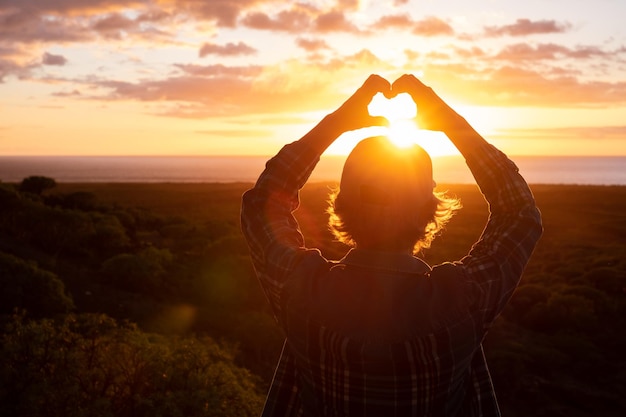 Foto vista trasera de una anciana con sombrero y ropa informal haciendo forma de corazón con las manos mientras mira la dorada luz del sol sobre el mar amor emoción y belleza en la naturaleza