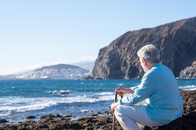 Vista trasera de un anciano de pelo blanco sosteniendo un bastón para caminar sentado en la playa mirando el horizonte sobre el agua