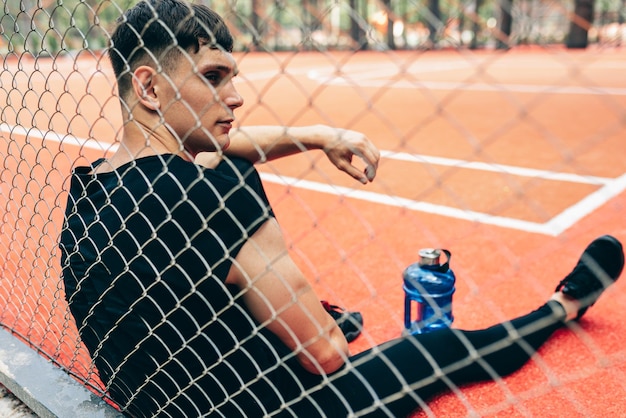 Vista trasera del apuesto joven fitness descansando después del entrenamiento con una botella azul de agua sentado en el campo de deportes Hombre sano tomando un descanso después de hacer ejercicio al aire libre