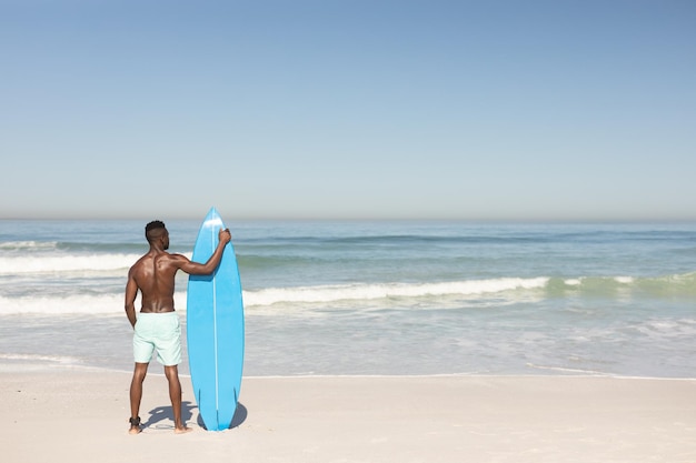 Una vista trasera de un atractivo afroamericano disfrutando del tiempo libre en la playa en un día soleado, sonriendo, divirtiéndose, de pie con su tabla de surf, el sol brillando sobre él.