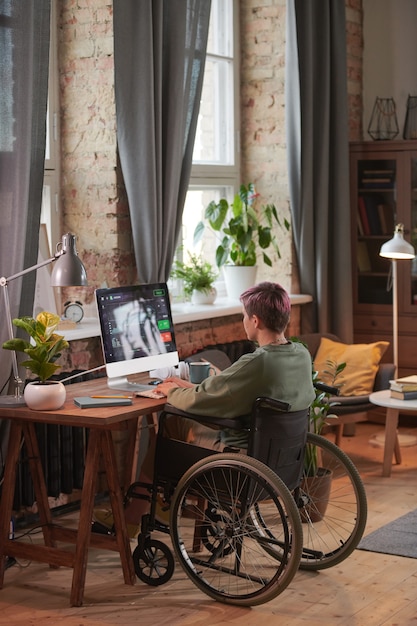 Vista trasera del autónomo discapacitado sentado en la mesa frente al monitor de la computadora y trabajando en línea en casa