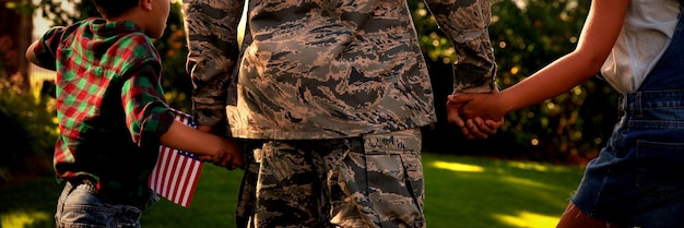 Vista trasera de cerca de un joven soldado adulto de raza mixta en el jardín fuera de su casa, tomándose de la mano con su hijo pequeño y su hija, su hijo sosteniendo una bandera estadounidense