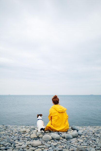 Vista trasera de una chica con impermeable amarillo sentada con un perro en una playa de guijarros mirando al mar