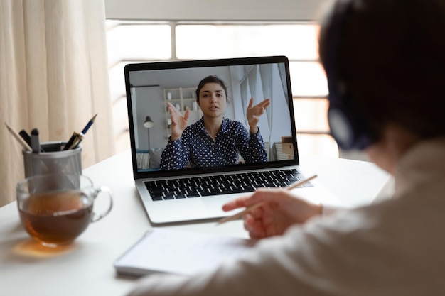 Vista trasera de la chica del milenio con auriculares escuchar ver capacitación en línea o curso en la computadora portátil empleada femenina hacer notas involucrada en conferencia de webcam o sesión informativa con colega tener reunión web en