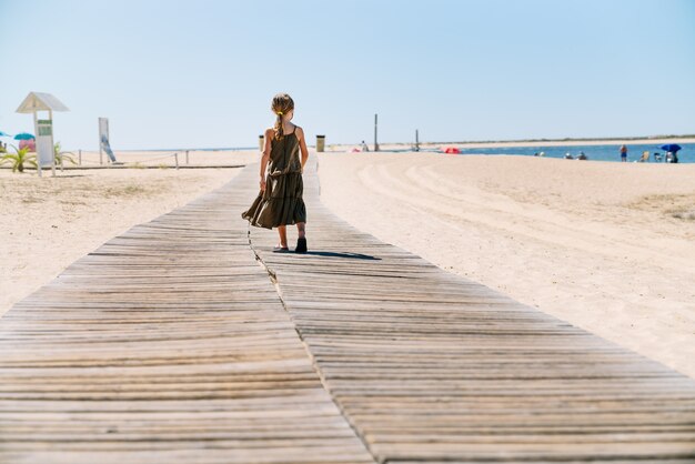 Vista trasera chica rubia con mascarilla de vacaciones caminando por un sendero en la arena de una playa con un vestido verde en medio de una pandemia de coronavirus