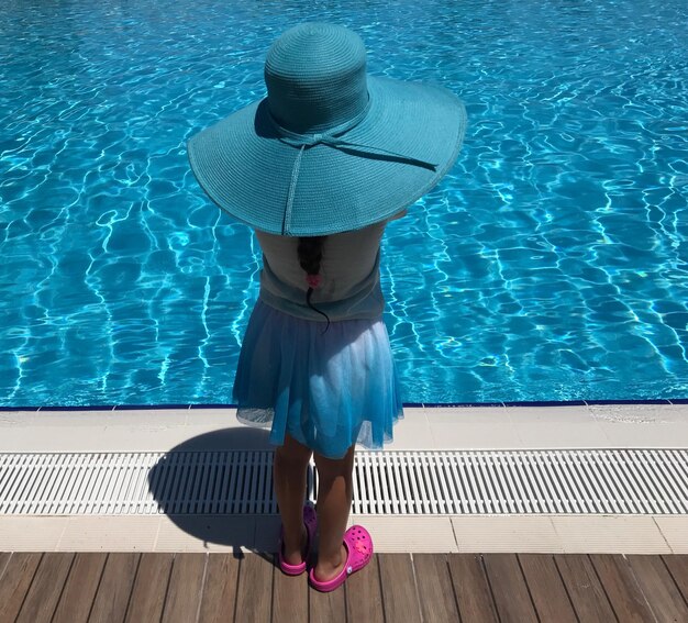 Vista trasera de una chica con un sombrero azul de pie junto a la piscina en un día soleado