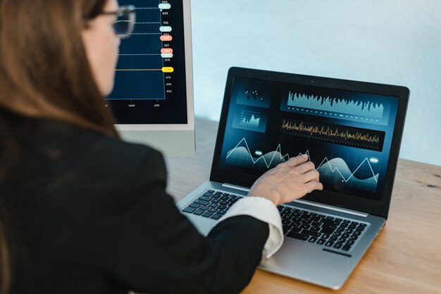 Vista trasera de una comerciante de cultivos que señala la pantalla de un netbook con gráficos financieros mientras trabaja en línea en la mesa