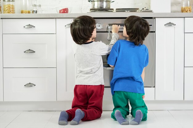 Vista trasera de curiosos niños hispánicos, gemelos viendo pasteles horneados en el horno, agachados en la cocina. Niños, concepto de cocina