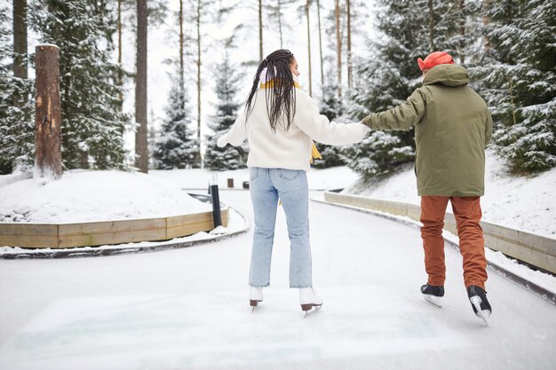 Vista trasera de dos amigos en ropa de abrigo tomados de la mano patinando juntos en el parque en día de invierno