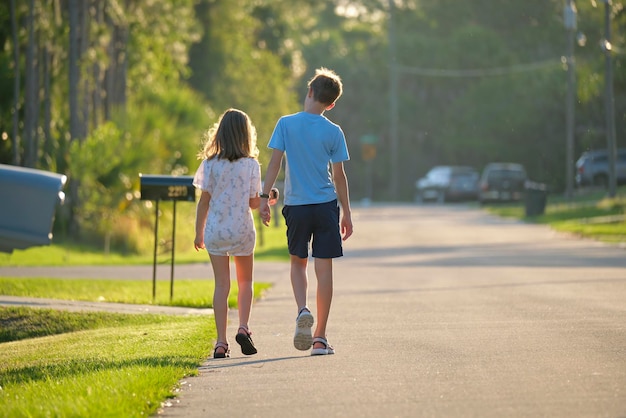 Vista trasera de dos niños adolescentes, una niña y un niño, un hermano y una hermana, caminando juntos por una calle suburbana en una brillante noche soleada.