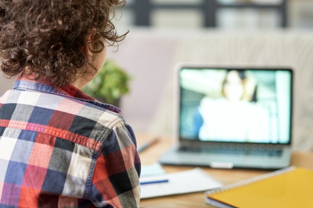 Vista trasera de la educación en línea del niño de la escuela latina tomando notas con lección en línea usando la computadora portátil