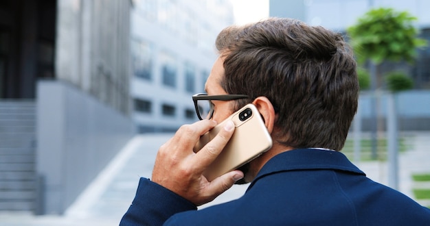 Vista trasera del elegante empresario hablando por teléfono celular y caminando en las calles de la ciudad. Hombre guapo hablando por teléfono inteligente en la ciudad. Vista trasera.