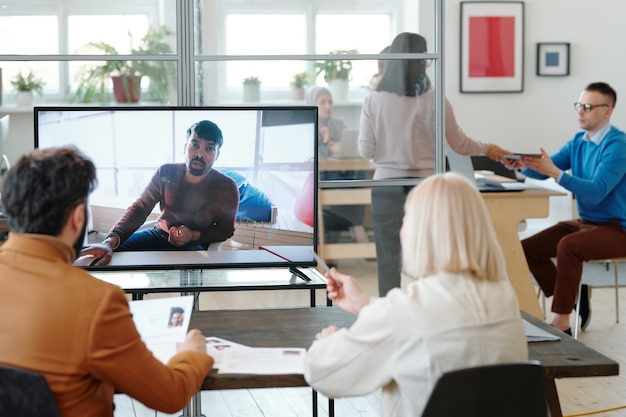 Vista trasera de especialistas en recursos humanos sentados en la mesa y viendo el currículum mientras habla con el solicitante de empleo a través de la aplicación de telecomunicaciones en la computadora