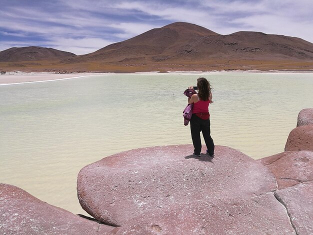 Vista trasera de una excursionista fotografiando mientras está de pie en una formación rocosa junto al lago