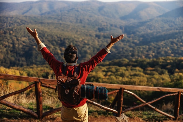 Vista trasera del explorador con las manos levantadas. Es un hermoso día soleado de otoño. Hombre con mochila y manta en la espalda. Libertad.