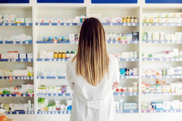 Vista trasera de una farmacéutica mirando estantes con medicamentos y drogas