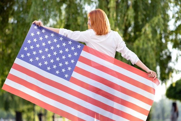 Vista trasera de la feliz joven pelirroja posando con la bandera nacional de Estados Unidos de pie al aire libre en el parque de verano. Chica positiva celebrando el día de la independencia de Estados Unidos.