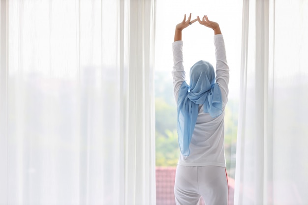 Vista trasera hermosa mujer musulmana asiática vistiendo ropa de dormir blanca, estirando los brazos después de levantarse por la mañana al amanecer. linda mujer joven con hijab azul de pie y relajante mientras mira lejos