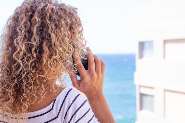 Vista trasera de la hermosa mujer rizada mientras usa el teléfono. Mar azul de fondo. Modelo femenino con espacio de copia