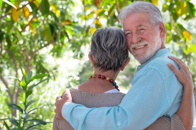 Vista trasera de una hermosa pareja de ancianos caminando por el bosque tiernamente abrazada mientras el hombre mira a la cámara Los abuelos ancianos sonrientes disfrutan de un estilo de vida saludable en el parque público