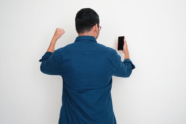 Vista trasera del hombre adulto apretando el puño mostrando emoción al mirar el teléfono que sostiene