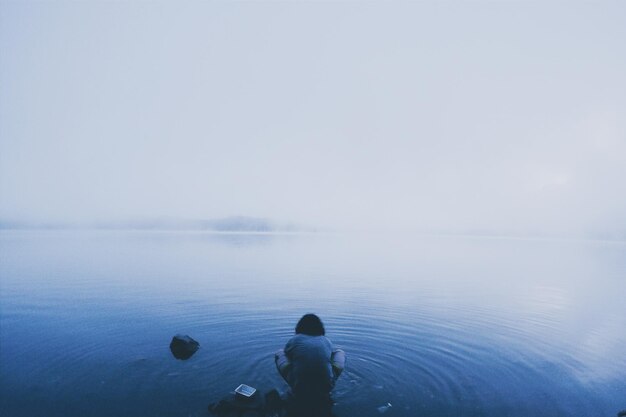 Foto vista trasera de un hombre agachado en la orilla del lago durante el tiempo de niebla