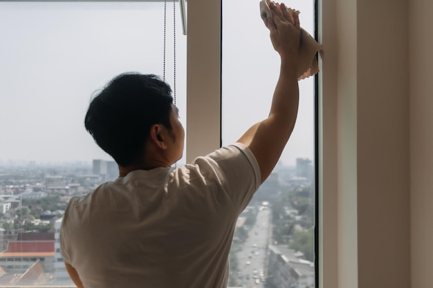 Vista trasera de un hombre asiático tailandés limpiando el vidrio de la ventana en un apartamento con vista a la ciudad.