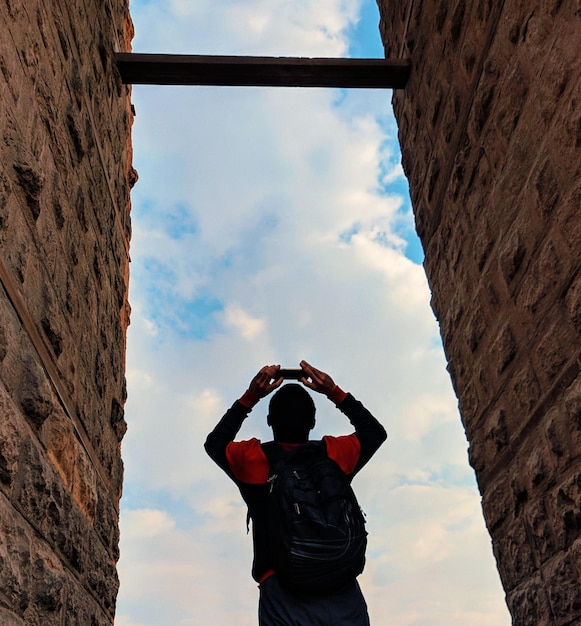 Foto vista trasera de un hombre fotografiando por una pared de piedra contra un cielo nublado