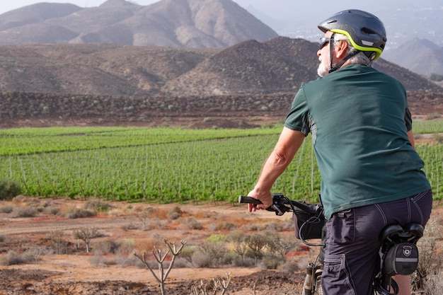 Vista trasera del hombre mayor con casco en su bicicleta eléctrica al aire libre entre viñedos