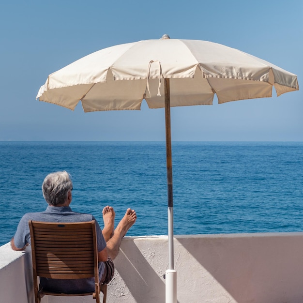 Vista trasera del hombre mirando al mar mientras está sentado en una silla con una sombrilla en la terraza del edificio