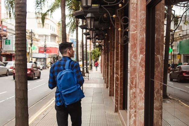 Vista trasera de un hombre con mochila y auriculares inalámbricos caminando por las calles de la ciudad.