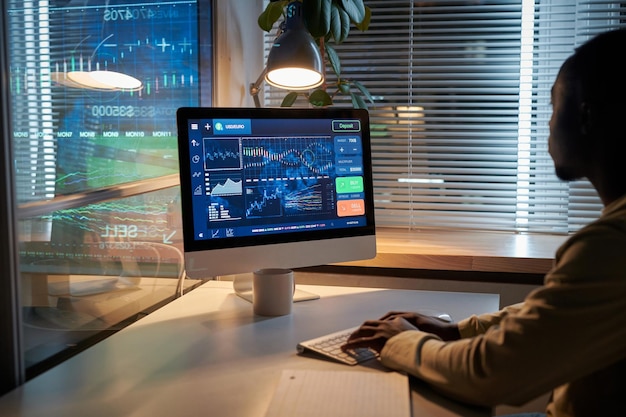 Vista trasera de un hombre de negocios africano mirando el monitor de la computadora y trabajando con software en la mesa de la oficina durante la fecha límite