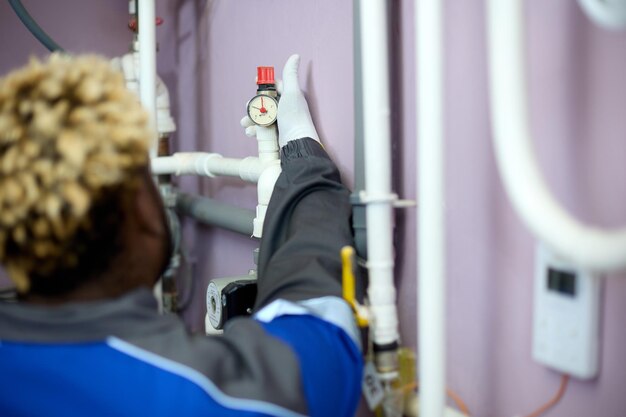 Vista trasera de un hombre negro con overol y guantes blancos que revisa el sensor de presión en la sala de calderas Concepto de mantenimiento de alta calidad de equipos técnicos complejos