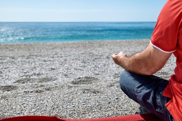 Foto vista trasera de un hombre con ropa casual sentado en postura de loto en una playa de guijarros y meditando en un día soleado