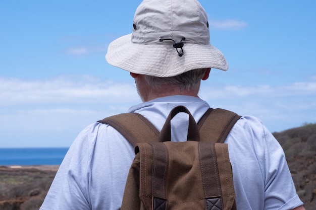 Vista trasera del hombre senior con sombrero disfrutando de senderismo al aire libre, mirando al horizonte sobre el mar