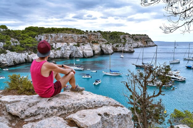 Vista trasera de un hombre sentado en una roca mirando al mar contra el cielo