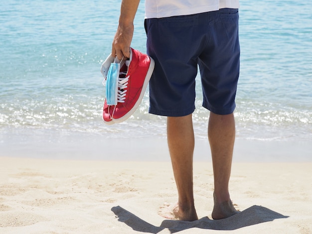 Vista trasera del hombre tiene máscara médica, zapatillas rojas en Sandy Tropical Beach.